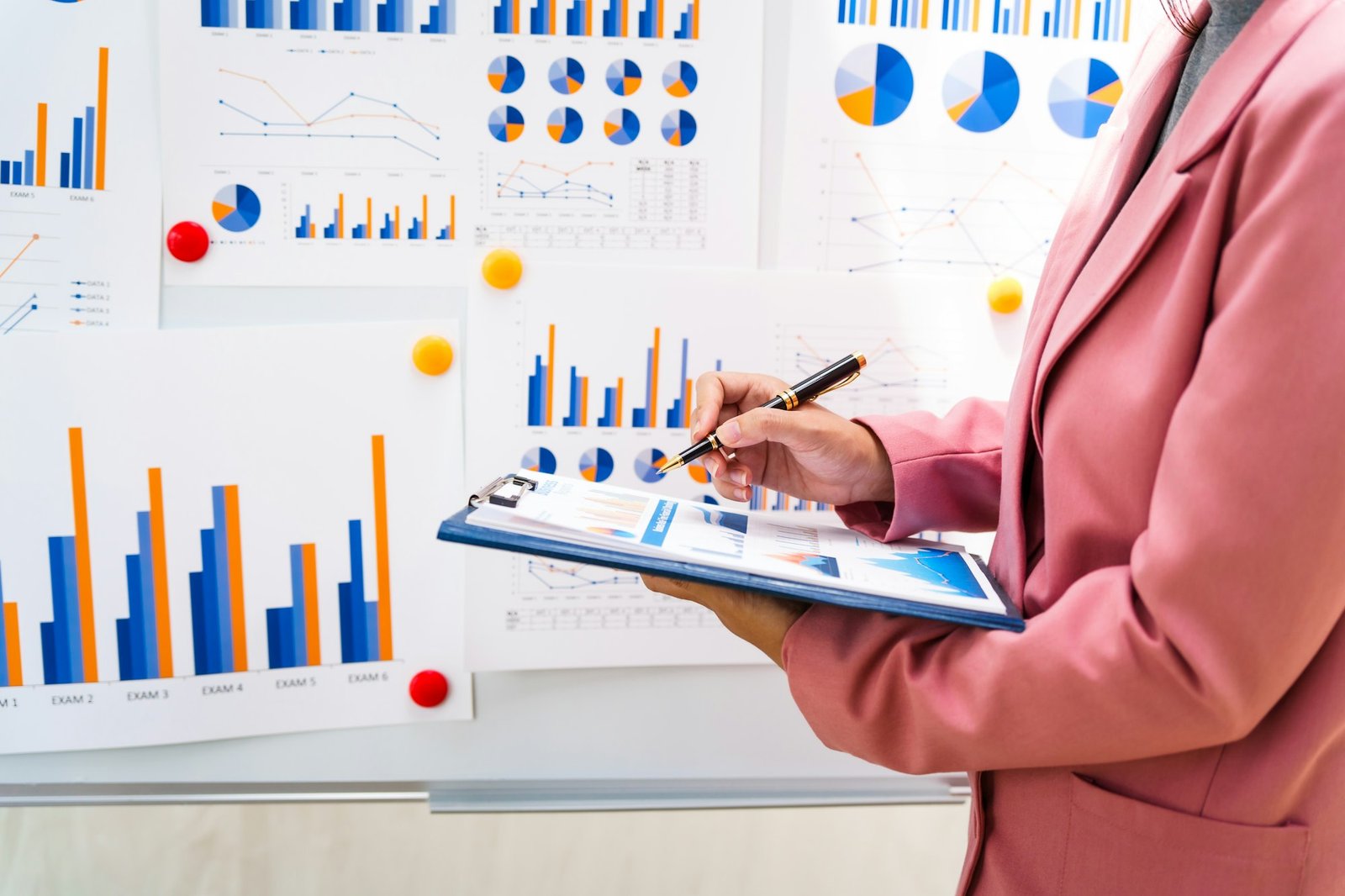 A businesswoman in a pink suit works at a desk with a whiteboard in the background.